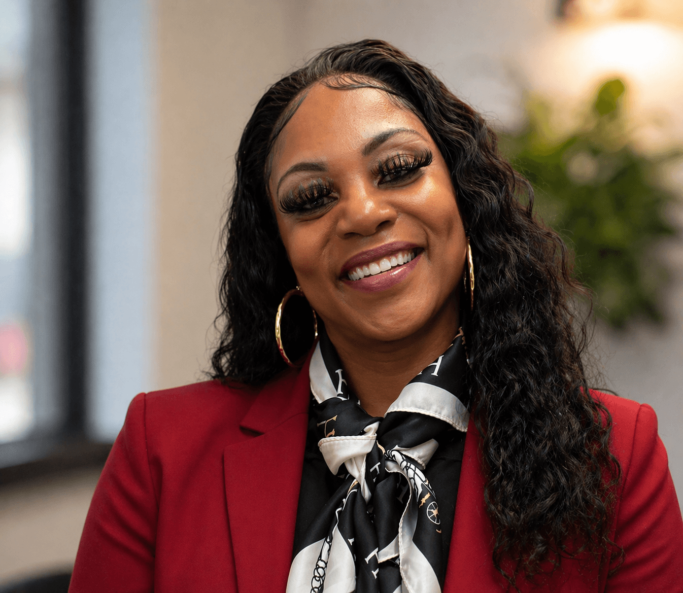 Smiling Black woman with long dark curls, wearing a red blazer and patterned silk scarf.